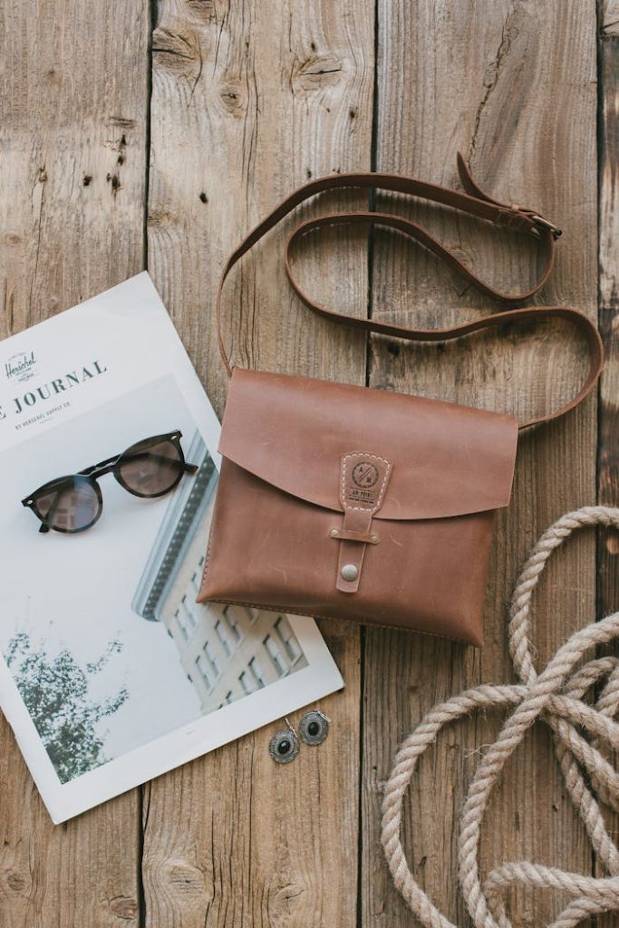 A stylish flat lay featuring a leather sling bag, rope, sunglasses, and a magazine on a rustic wooden table.